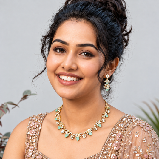 A smiling woman with dark hair styled in an updo, wearing a gold-toned floral necklace and matching drop earrings with mint green stones, paired with a heavily embellished beige gown against a soft gray background.