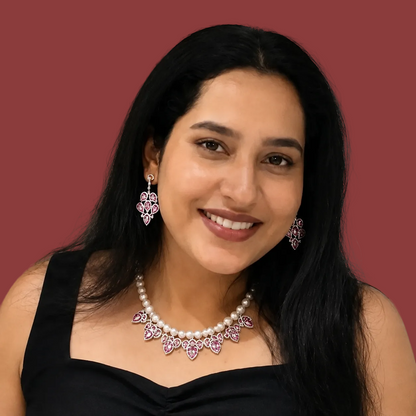 A woman with long black hair smiling, wearing a black top and a pearl necklace with pink gemstone floral accents, along with matching pink and silver earrings, against a neutral indoor background.