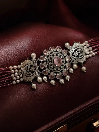 An ornate silver-toned traditional Indian bridal forehead band (tilak) decorated with pink gemstones, white pearls, and intricate filigree, resting on a dark burgundy leather surface.