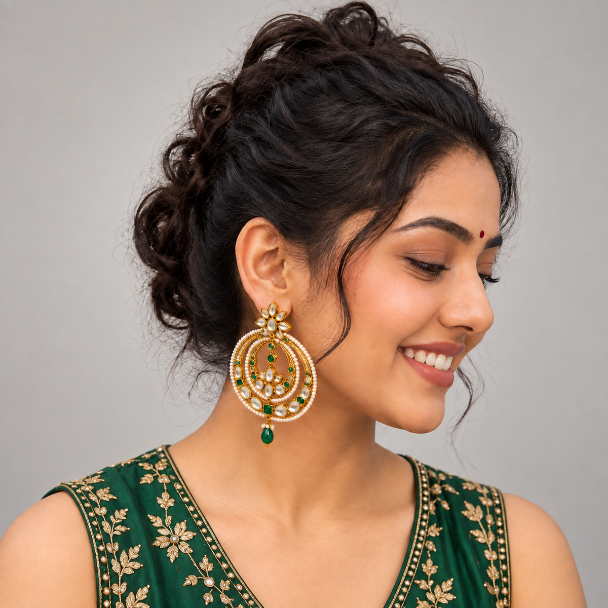 A woman wearing a large gold-toned traditional Indian jhumka earring with green stone accents, shown in a close-up portrait against a plain gray background.