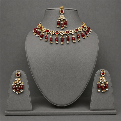 Gold-plated Indian jewelry set featuring a necklace, matching earrings, and a topkara with deep red stones and white crystal accents, displayed on a gray velvet stand.