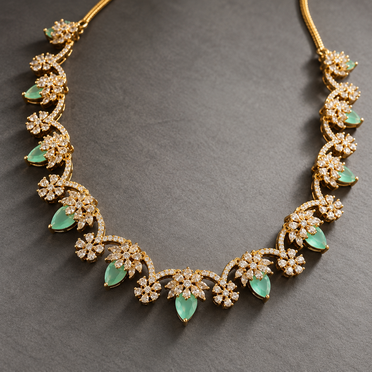 Gold floral filigree necklace with green teardrop emerald-cut stones and pavé-set clear stones, displayed on a dark gray textured background.