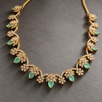 Gold floral filigree necklace with green teardrop emerald-cut stones and pavé-set clear stones, displayed on a dark gray textured background.