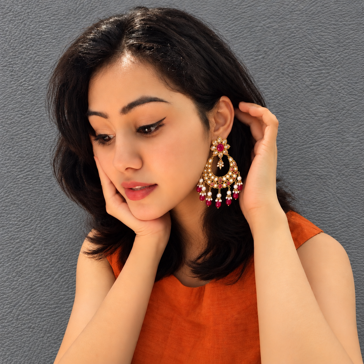 A woman wearing a large, ornate gold-toned chandelier earring with red and white bead details, posing against a textured gray wall.