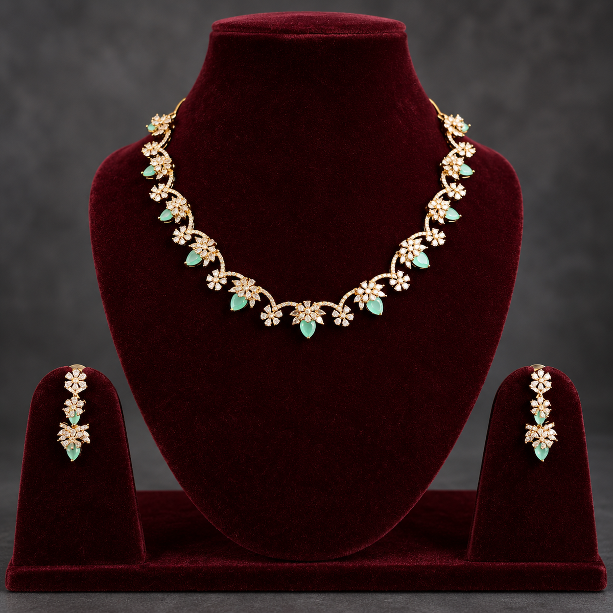 Gold floral jewelry set with mint green stones, displayed on a dark red velvet necklace bust and matching stand, with matching drop earrings on side stands against a dark gray background.