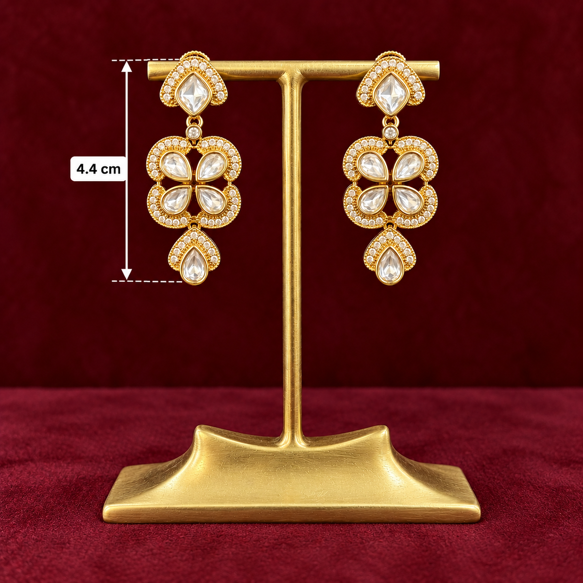 Gold-plated statement dangle earrings with clear crystals, displayed on a gold stand against a dark red background, with a size marker indicating 4.4 cm height.