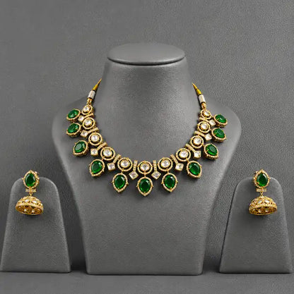 Gold-plated jewelry set featuring a statement necklace and matching jhumka earrings with green stones and clear accent stones, displayed on a gray mannequin bust.