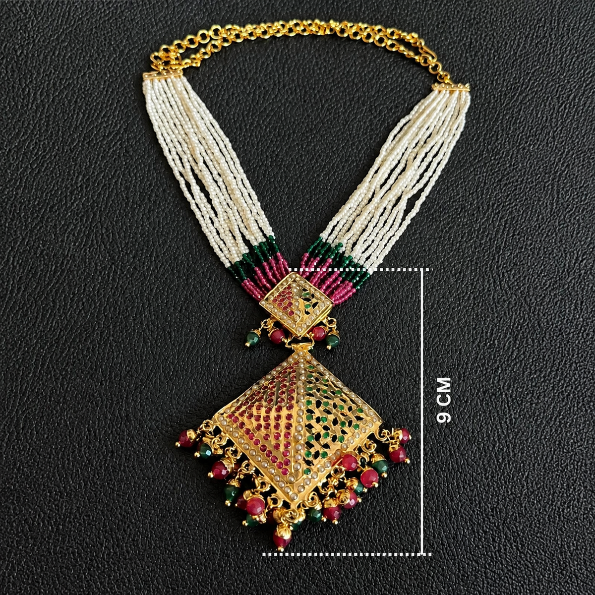 An ornate gold-toned statement necklace with multi-strand cream bead strands, red and green stone accents, and a large diamond-shaped pendant, shown on a black textured background with a 9 cm size marker.