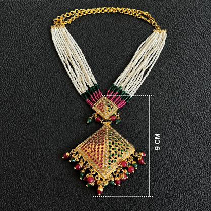 An ornate gold-toned statement necklace with multi-strand cream bead strands, red and green stone accents, and a large diamond-shaped pendant, shown on a black textured background with a 9 cm size marker.
