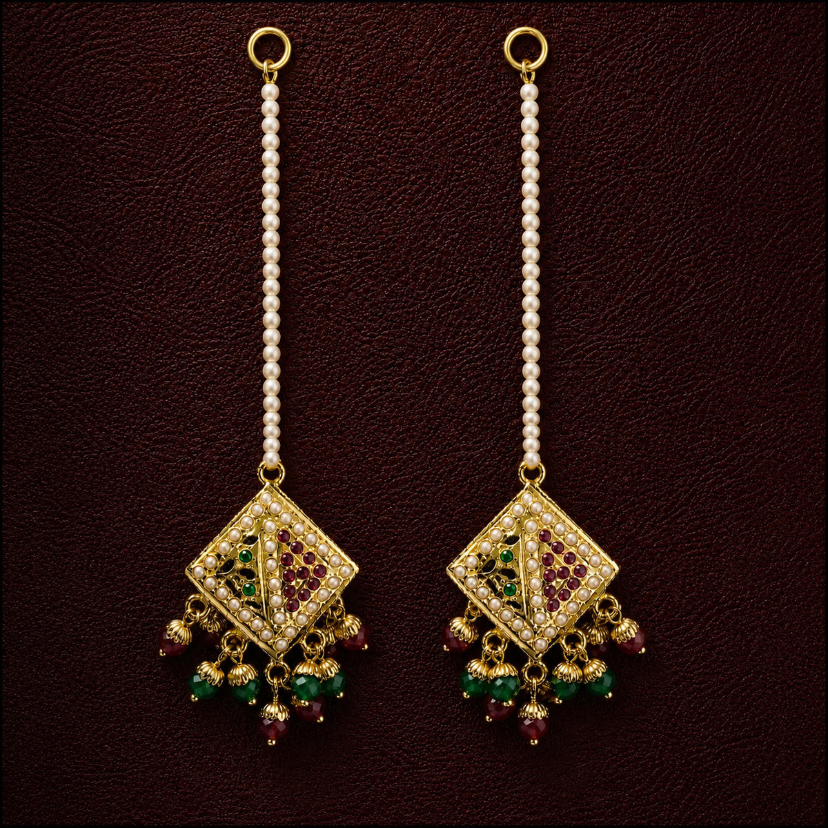 A pair of traditional Indian gold-plated dangle earrings with white pearl bead chains, featuring a diamond-shaped pendant adorned with red stones and hanging green and red bead drops, set against a dark brown textured background.