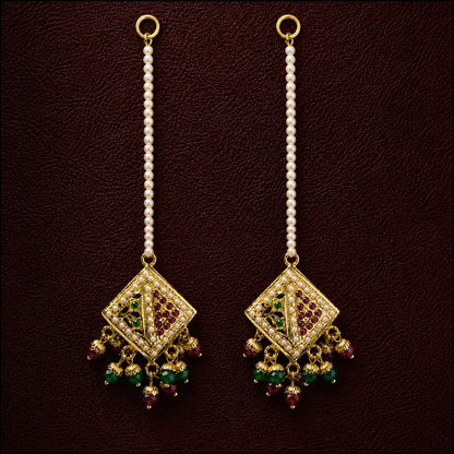 A pair of traditional Indian gold-plated dangle earrings with white pearl bead chains, featuring a diamond-shaped pendant adorned with red stones and hanging green and red bead drops, set against a dark brown textured background.