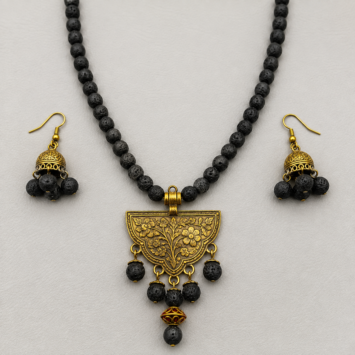 A jewelry set featuring a necklace and matching earrings made of dark black lava stones and ornate gold-toned metal, displayed on a light gray background.