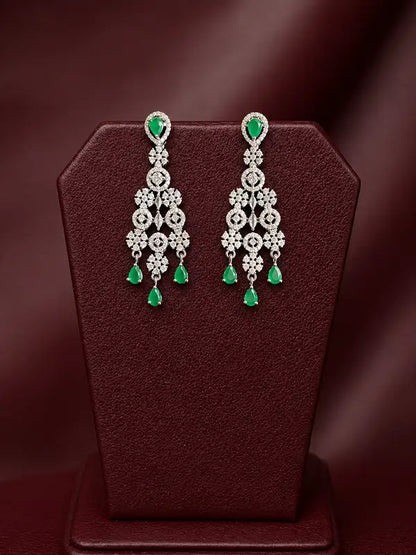 A pair of ornate silver-toned dangle earrings with green teardrop gemstones and floral silver motifs, displayed on a dark burgundy jewelry stand against a dark maroon background.