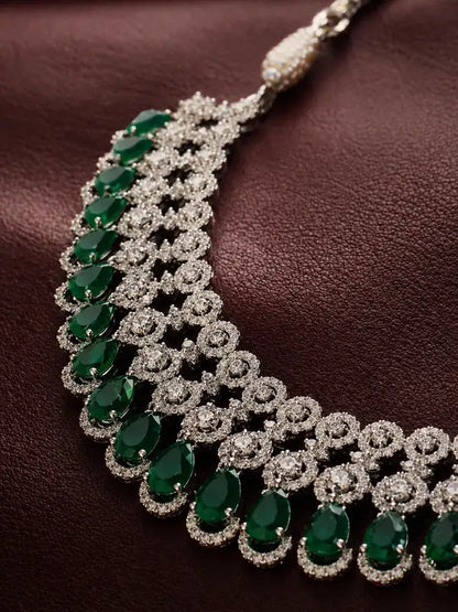 An ornate silver-toned statement necklace featuring rows of oval and teardrop-cut emerald green gemstones surrounded by pavé-set clear stones, displayed on a dark brown textured leather surface.