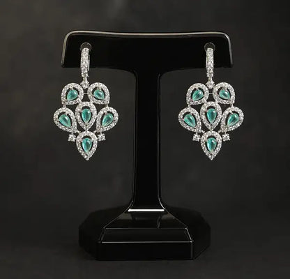 A pair of silver-toned drop earrings with teardrop-shaped green gemstones surrounded by small clear stones, displayed on a black T-shaped jewelry stand against a dark gray background.