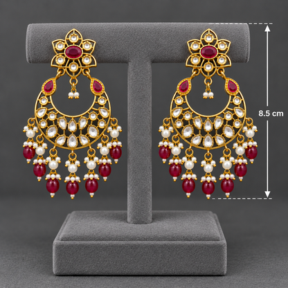 A pair of ornate gold-toned dangle earrings displayed on a gray jewelry stand against a dark gray background. The earrings feature red gemstone accents, white pearl-like beads, and clear stones, with a vertical measurement marker indicating 8.