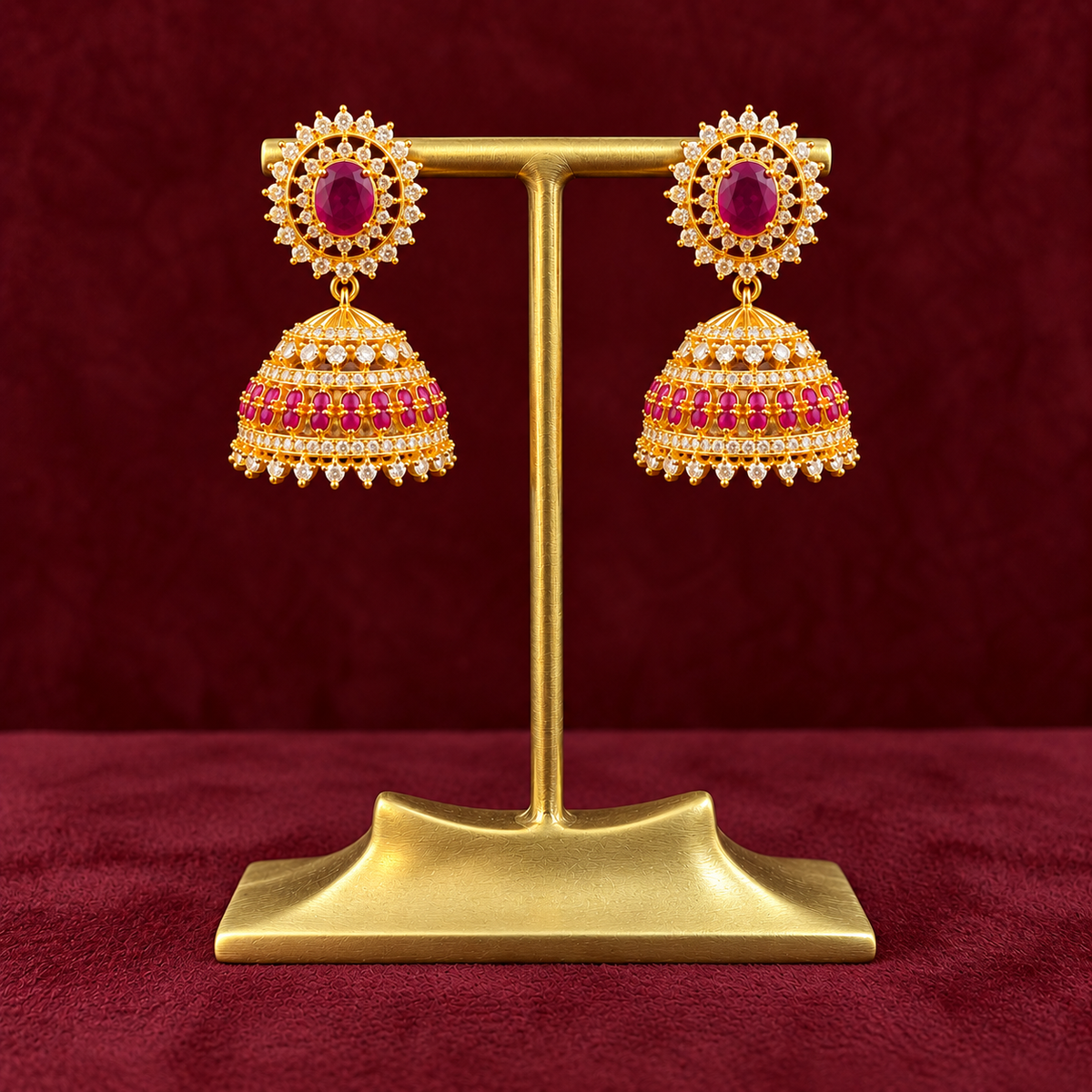 A pair of gold-toned traditional jhumka earrings with red gemstone centers and pink stone detailing, displayed on a gold stand against a dark red textured background.