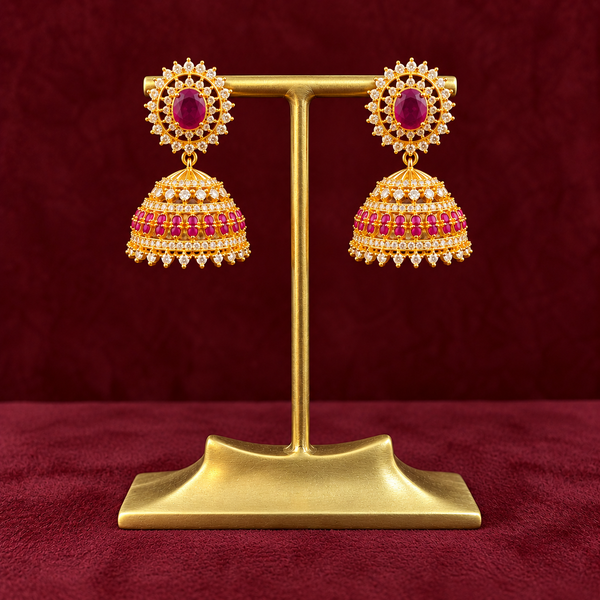 A pair of gold-toned traditional jhumka earrings with red gemstone centers and pink stone detailing, displayed on a gold stand against a dark red textured background.