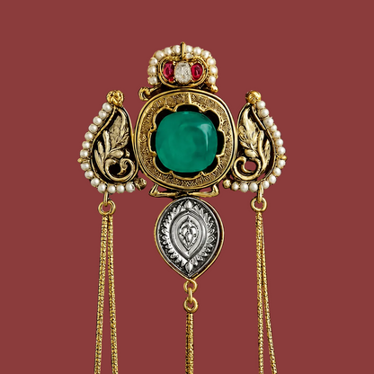 An ornate vintage-style brooch or pendant in gold-tone metal with a large green cabochon center stone, pearl-like white accents, a small red and clear stone crown detail, and dangling gold chains, displayed against a dark burgundy background.