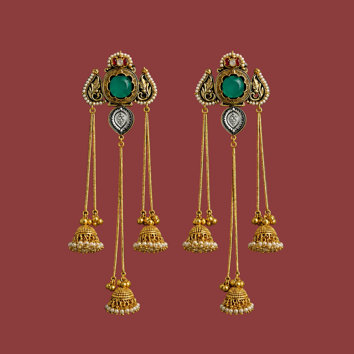 A pair of ornate gold-toned traditional dangle earrings displayed on a gray presentation box. Each earring features a decorative top section with a green oval stone, a small red stone, a white heart-shaped stone, and multiple dangling bell-shaped charms embellished with small white stones.
