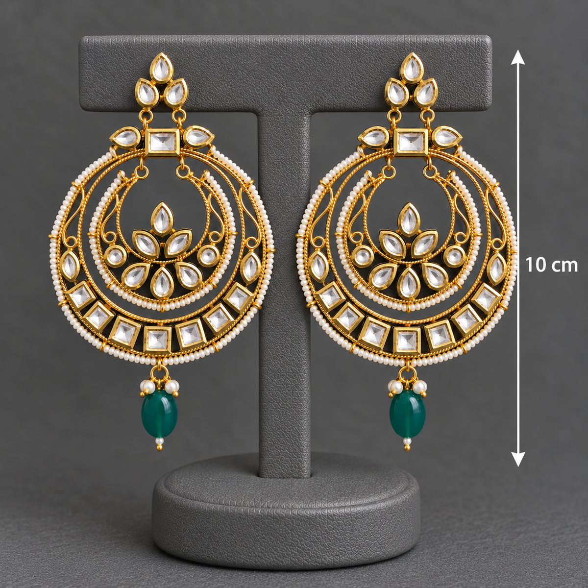 A pair of ornate gold-toned Indian-style dangle earrings with white stones and green teardrop drops, displayed on a gray T-stand with a 10 cm height marker against a dark gray background.