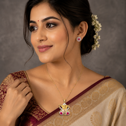 A woman wearing a gold pendant necklace with a Ganesha design and matching stud earrings, styled with a traditional Indian saree against a neutral gray background.