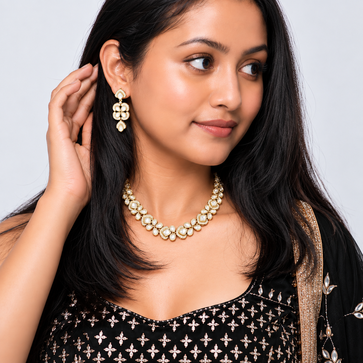 A woman modeling a gold-toned traditional jewelry set featuring a matching necklace and dangle earrings with white stones, set against a plain light gray background.