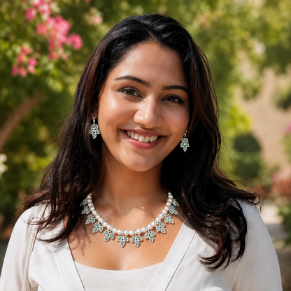 A smiling woman with long dark hair wearing a pearl necklace and matching drop earrings with silver-toned floral motifs, photographed outdoors against a blurred green and pink garden background.