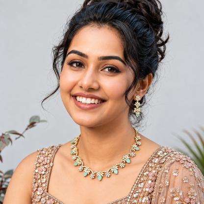 A smiling woman with dark hair styled in an updo, wearing a gold-toned floral necklace and matching drop earrings with mint green stones, paired with a heavily embellished beige gown against a soft gray background.